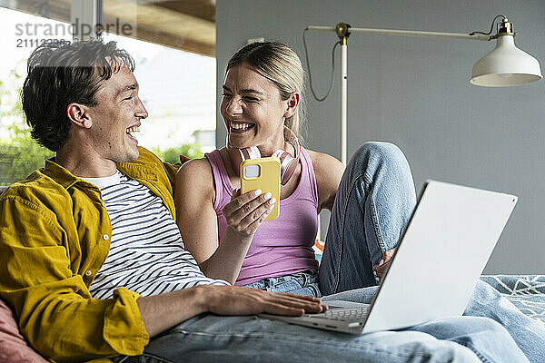 Cheerful woman holding smart phone spending leisure time with boyfriend at home