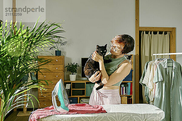 Woman holding black cat standing near ironing board at home