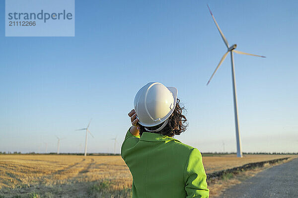 Engineer wearing hard hat and looking at wind turbine in farm