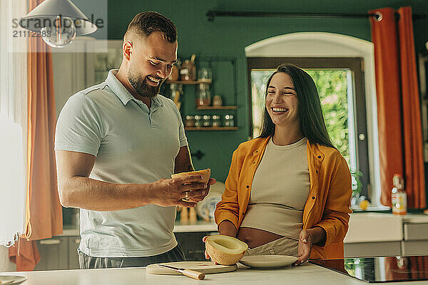 Happy pregnant woman with husband holding muskmelon in kitchen at home