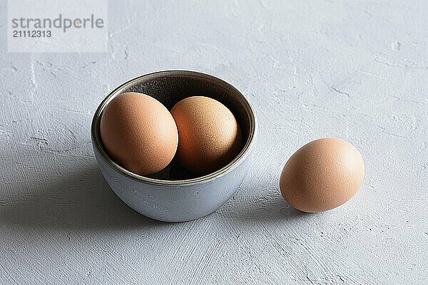 Studio still life of a bowl with three brown eggs and a single egg on a textured surface.