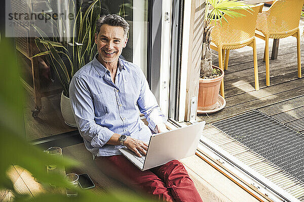 Portrait of man smiling while using laptop by open balcony