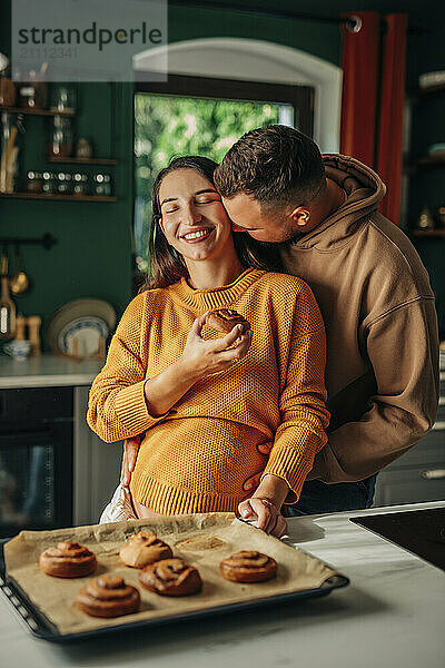 Affectionate husband with pregnant wife holding cinnamon bun in domestic kitchen