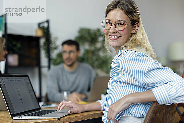 Happy businesswoman wearing eyeglasses looking over shoulder and sitting with laptop in office