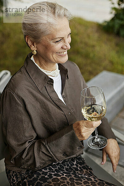 Happy senior woman sitting and holding glass of white wine at restaurant