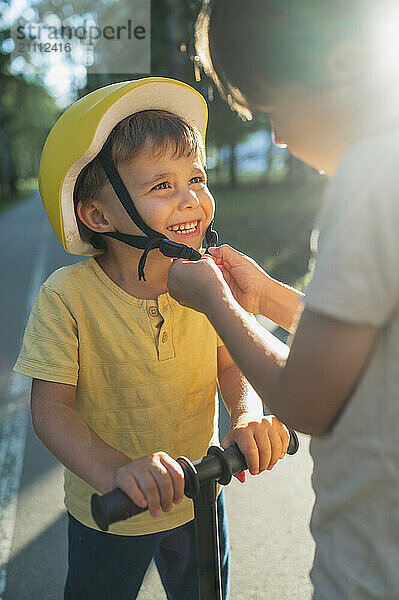 Happy boy in push scooter wearing helmet with help of brother on road