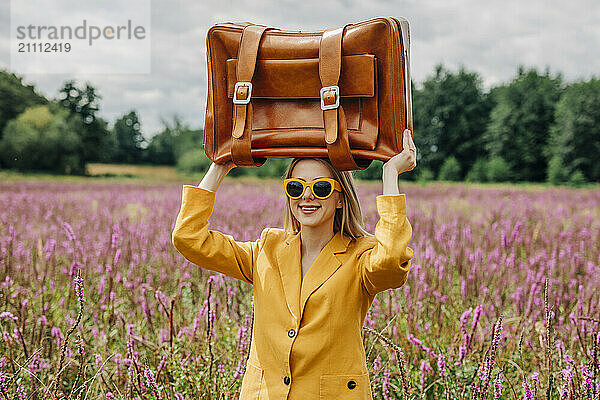 Playful woman holding brown suitcase on head and standing in field