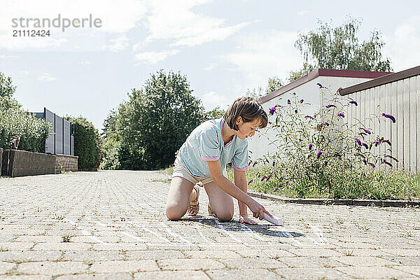 Girl kneeling and drawing with chalk on paving stone at sunny day