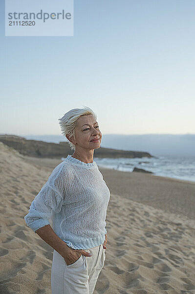 Thoughtful woman with hands in pockets standing at beach