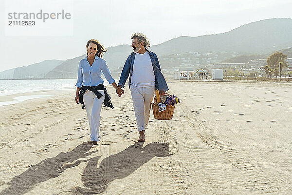 Happy senior man holding basket and walking with woman at beach