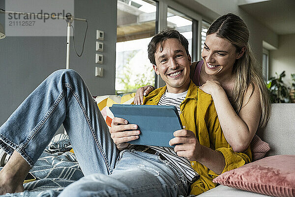 Smiling woman leaning on boyfriend holding tablet PC on sofa at home