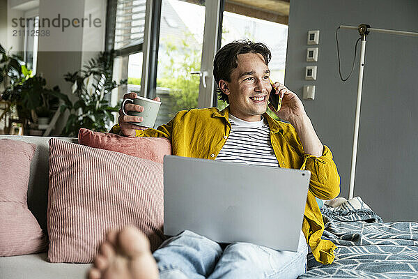 Smiling man holding coffee cup and talking through smart phone on sofa at home