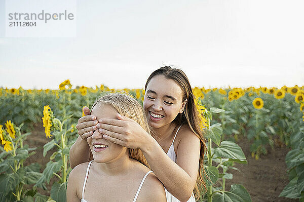 Happy woman covering eyes of sister in sunflower field