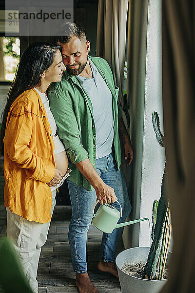 Smiling man watering plant with pregnant wife at home