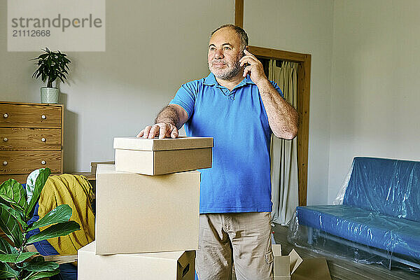 Senior man talking on smart phone and standing near boxes in new apartment