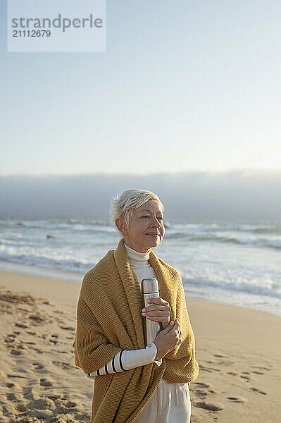 Smiling woman wrapped in shawl standing on beach near sea