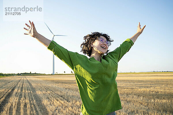 Cheerful young woman jumping in field near wind turbine generator