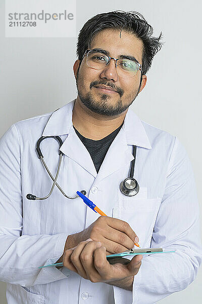 Smiling young doctor holding pen and clipboard against white background