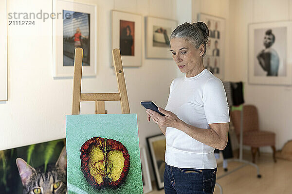 Artist using smart phone and standing near painting in studio