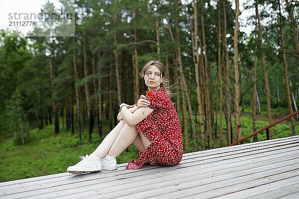 Woman in red dress sitting on wooden patio near forest