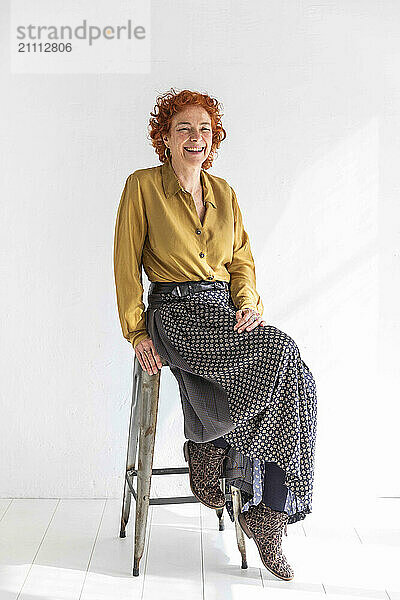 Happy woman sitting on stool in front of white wall