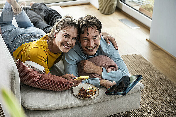 Smiling woman with arm around boyfriend lying on sofa at home