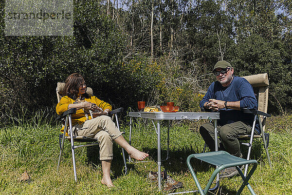 Happy senior couple spending leisure time sitting on camping chairs near trees