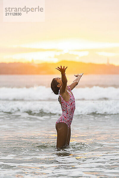 Girl in swimsuit standing in water with arms outstretched at beach