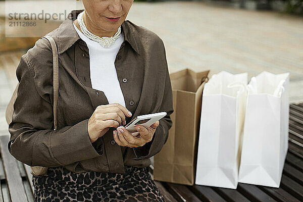 Mature woman using smart phone and sitting with shopping bags on bench at street