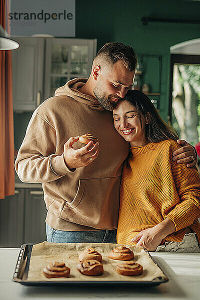 Young man holding cinnamon bun with arm around pregnant wife in kitchen