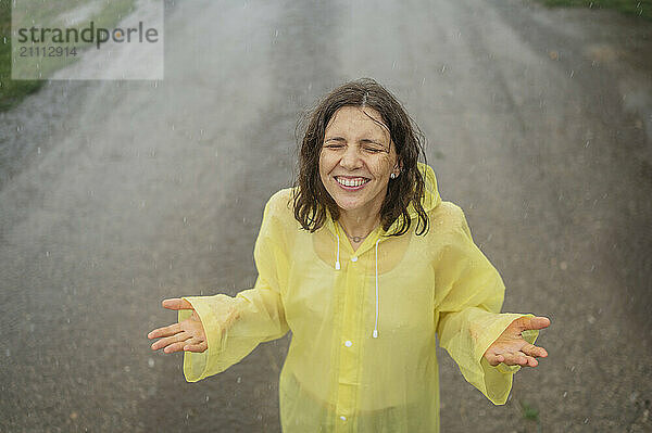 Happy woman in raincoat standing with eyes closed and enjoying rain on road