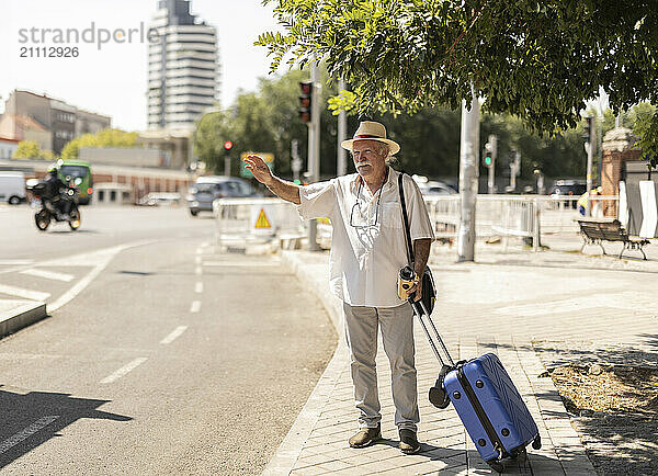 Senior man standing with suitcase and hailing ride on sunny day