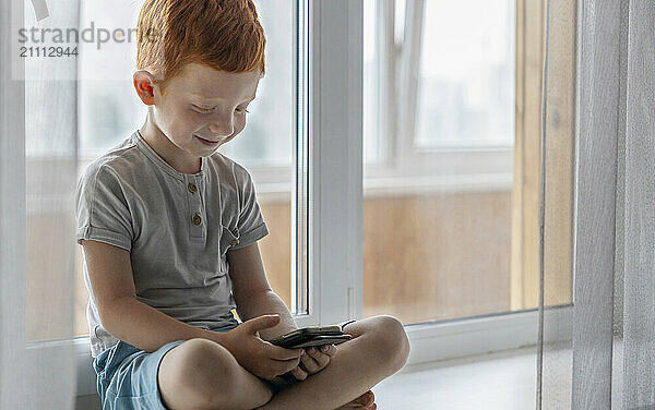 Redhead boy using smart phone and sitting cross legged on window sill