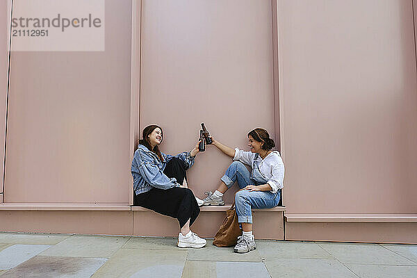 Happy couple toasting with beer bottles and sitting in front of pink wall