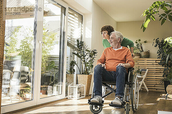 Senior woman pushing husband on wheelchair near glass door at home