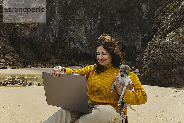 Senior freelancer carrying dog and holding laptop at beach