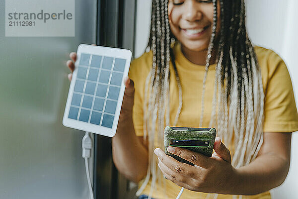 Smiling girl charging smart phone with solar panel equipment at home