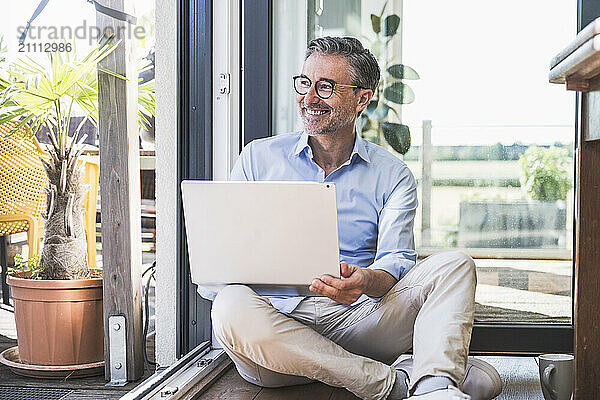 Smiling businessman with laptop sitting on floor at home office