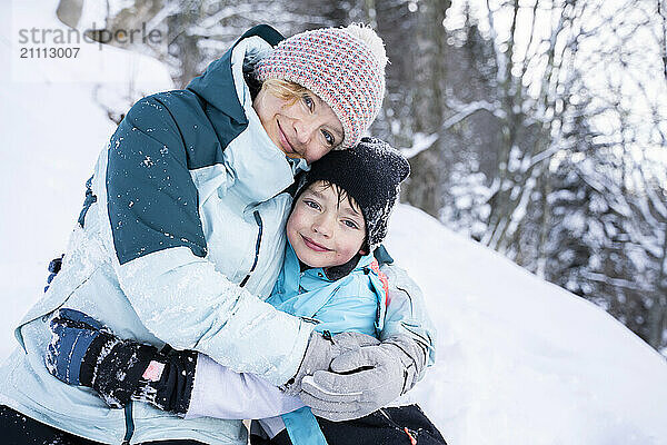 Smiling mother embracing son sitting on snow at winter