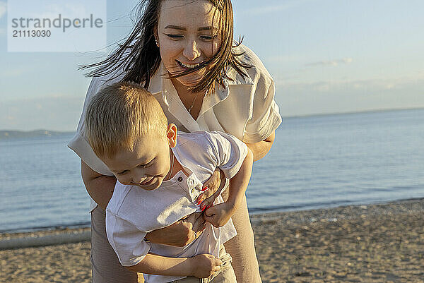 A dark-haired happy woman in a white shirt with her son on the beach.