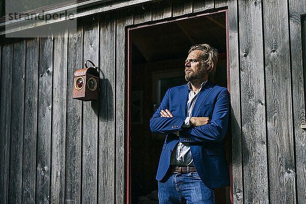 Thoughtful businessman standing with arms crossed at doorway of log cabin