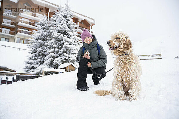 Happy woman crouching with doodle dog on snow in winter