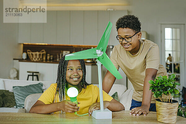 Happy brother with sister powering light bulb through wind turbine model in living room at home