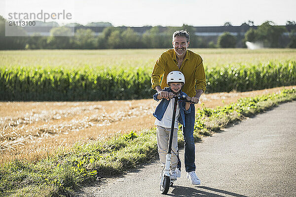 Smiling grandson riding push scooter with grandfather on road