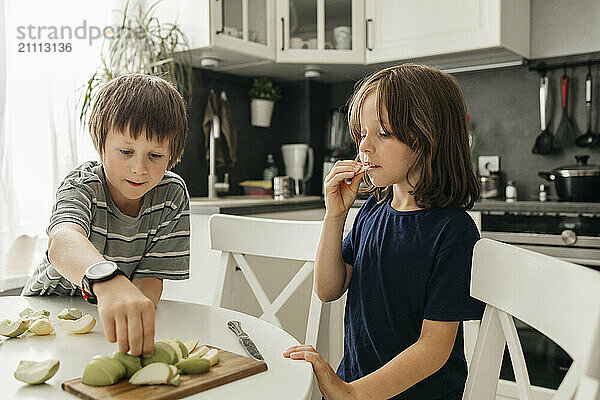 Siblings sitting near table and eating sliced apple in kitchen