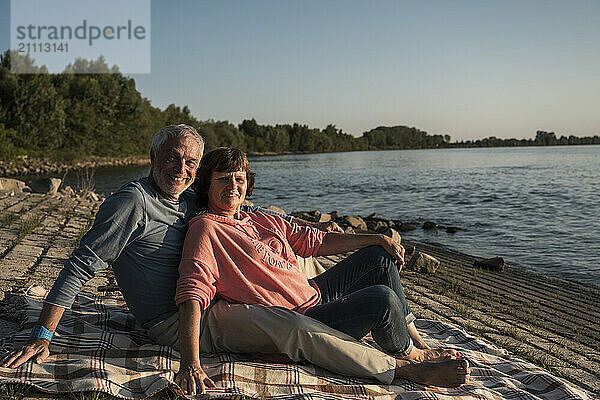 Happy senior couple sitting on picnic blanket at riverbank