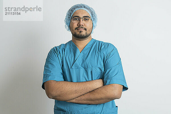 Young doctor in medical scrubs with arms crossed against white background