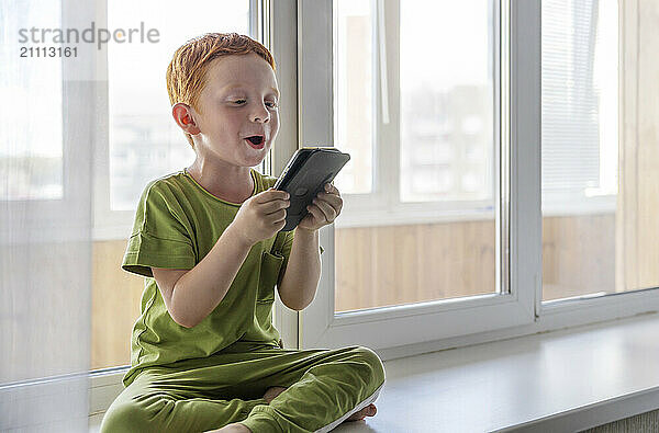 Cute boy with smart phone sitting on window sill and enjoying at home