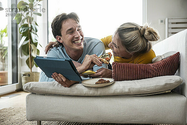 Cheerful man feeding pizza to girlfriend lying on sofa at home