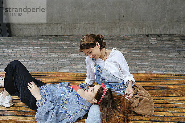 Young woman relaxing on lap of girlfriend sitting on bench in front of wall